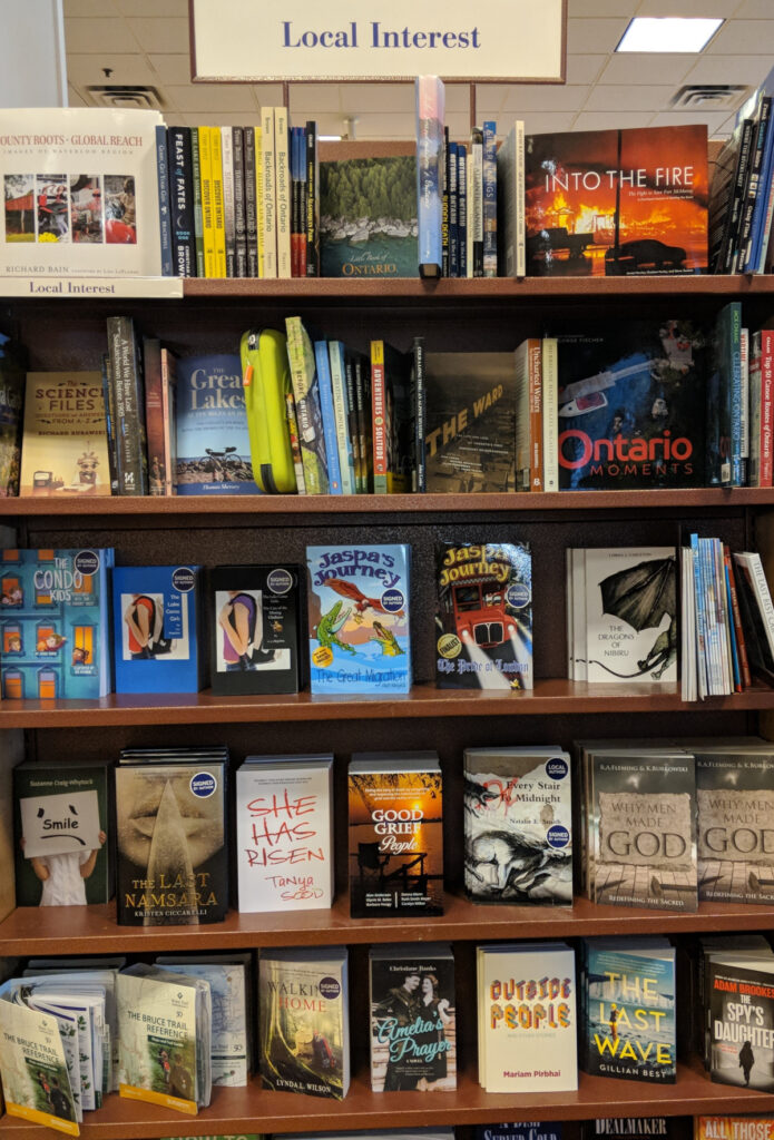 A crowded shelf in a bookstore headed "local interest" includes among the titles a copy of Mariam Pirbhai's Outside People (2017).